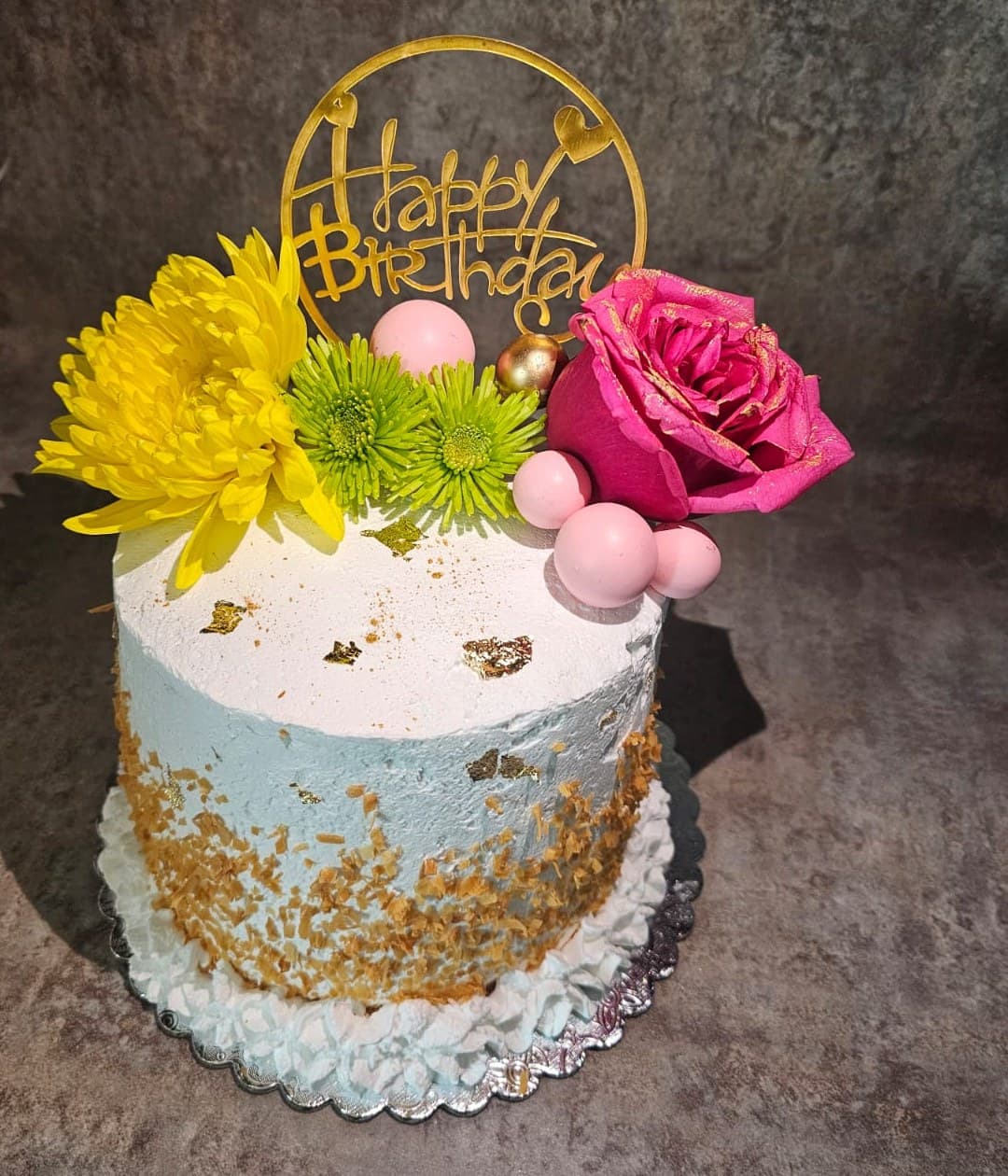 Birthday cake topped with a yellow chrysanthemum, green mums, a pink rose and a gold Happy Birthday ring.