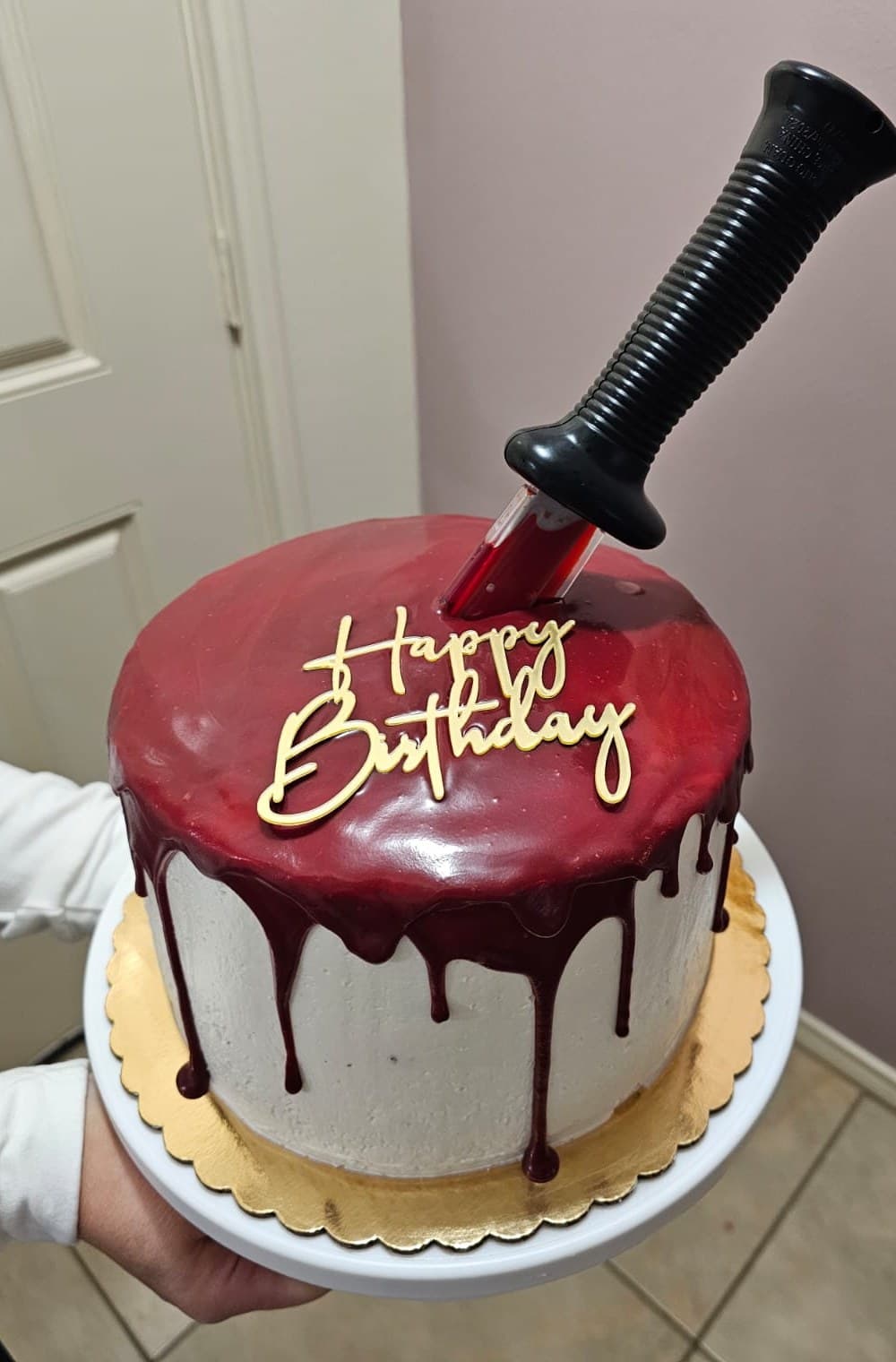 White birthday cake with a glossy red drip, a gold Happy Birthday topper and a prop knife on top.