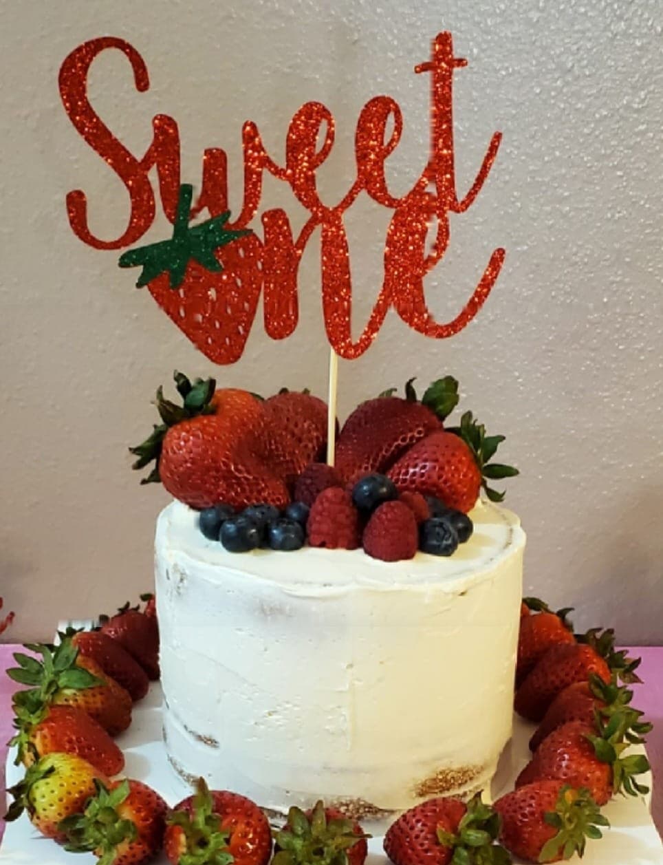 White first-birthday cake piled with strawberries, raspberries and blueberries, ringed with fresh strawberries, under a red glitter “Sweet One” topper.